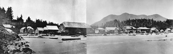 A view from the water of a sandy beach, with canoes and logs lying on the sand. Behind the sand is a long line of buildings and houses, backed by evergreen trees. At the right side of the picture, there are mountains behind the evergreens and two people are walking on the sand in the distance.