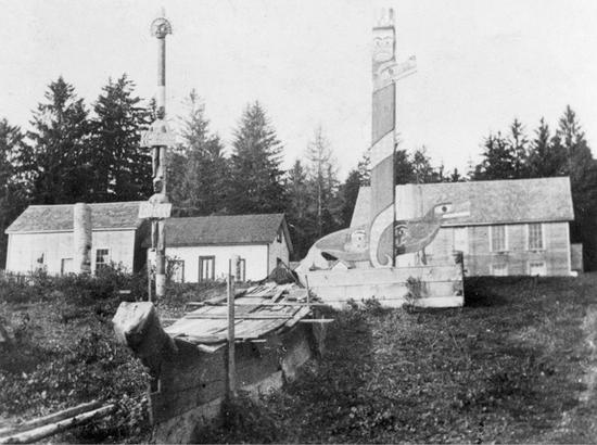 A canoe sealed shut with wood is lying on the ground in front of two carved poles. The left pole is carved in the shape of two people holding plaques, one standing on the other's shoulders, with a face at the top. The right pole is painted as if it's wrapped by a giant snake, with a face on top and a large bird at the bottom. There are three buildings behind the poles.