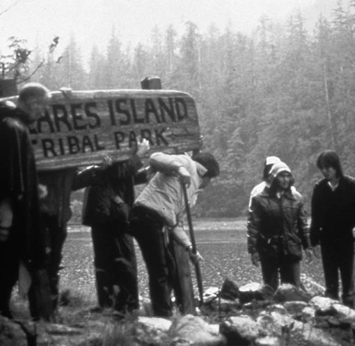 People installing a large wooden sign that reads 'Meares Island Tribal Park.' One person is digging with a shovel, and three more are holding the sign upright. Three more people are watching from the right side.