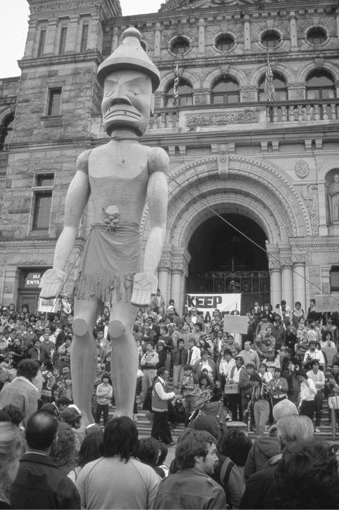 A large crowd of people is in front of a stone building. In the middle of the crowd is a giant wooden carved statue of a man. The statue has its palms upturned and is wearing a fringed cloth around its waist. Its hat is the same shape as the one worn by Maquinna.