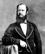 Light-skinned man with curly hair thinning at the top and a full beard, wearing a wide collared blazer on top of a white shirt and striped bow tie, leans one arm on a stand covered in cloth, looking slightly to the side