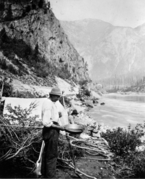 Dark-skinned man wearing a brimmed hat, loose shirt and long pants, holding a shallow pan and shovel looks down, standing in front of a tent. An older man with a beard is sitting against the tent, on the bank of a river in the mountains