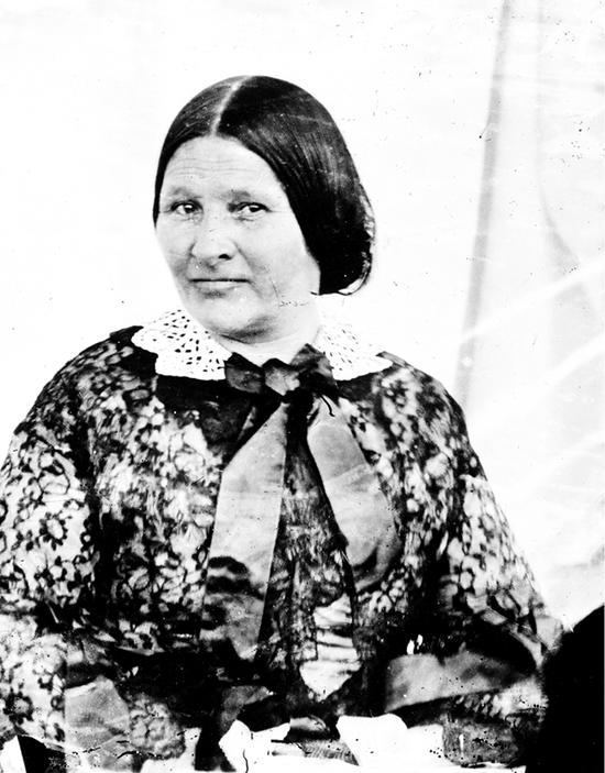 Middle-aged woman with long hair parted in the middle into a low bun wearing a floral dress with a lace collar tied with a thick ribbon, sitting and looking slightly to the side