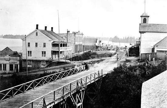A wooden bridge leads into a smooth road lined with two to three story buildings with glass windows, tiled roofs and chimneys, a river surrounded by trees borders the town