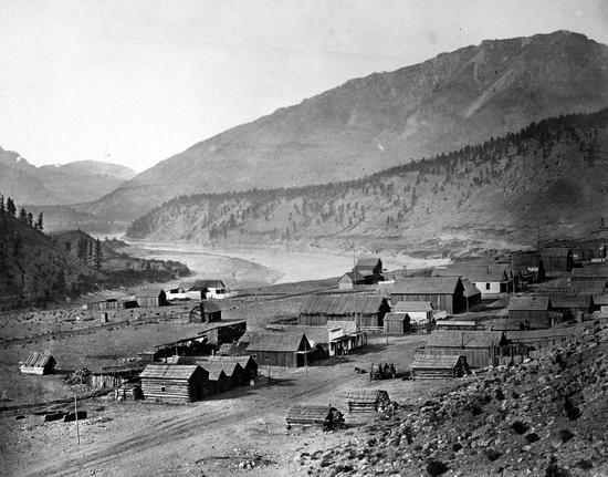 Three rows of wooden houses set near a river are separated by two dirt roads, surrounded by rocky mountains dotted with trees
