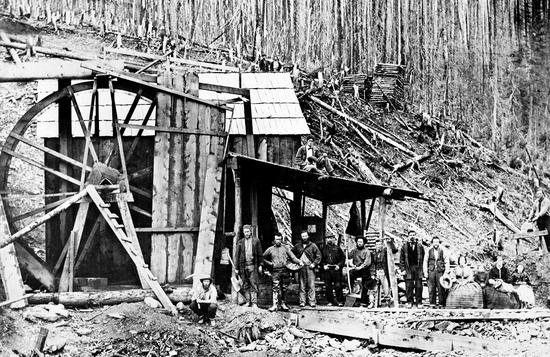 Nine men, one crouching and the rest standing, holding mining tools and hats, are to the right of four sitting females, all of them in front of a tall, simple wooden structure with a large wheel attached to the side. A man sits atop of a roofed area to the side of the building