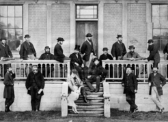 Securing pensions for British Columbia’s many civil servants , including members of the Legislative Assembly, pictured ca. 1870, was one of the preconditions of Confederation. From left to right on porch: W.A. Franklin, house messenger; Henry Holbrook; Wymond Ogilvy Hamley; Edward Graham Alston; Philip J. Hankin; Montague William Tyrwhitt-Drake; Joseph M. Trutch (sitting on porch at top of steps); Thomas L. Wood; Henry Maynard Ball; Edgar Dewdney; Arthur Thomas Bushby. From left to right in foreground: Amor de Cosmos; Henry Pering Pellew Crease; D.B. King (seated on steps, with dog); Thomas B. Humphreys; John Robson. Image C-06178 courtesy of the Royal BC Museum.