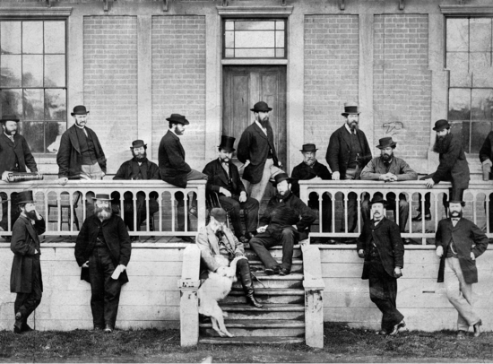Sixteen men wearing dark coats and brimmed hats and a pale dog are on the porch of a bricked building with glass windows and a carved, wooden railing