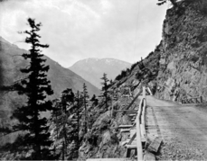 A wooden bridge connects rocky roads set into the mountainside, overlooking a steep drop to the side