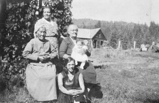 A woman stands in front of a leafy hedge with two older women seated, one with glasses and holding a baby girl, and a young woman with glasses sits on the grass, a wooden house and forest in the background
