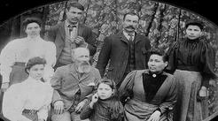Four women and a girl wearing dresses with puffy, loose shirts with high collars and heavy skirts, sit and stand around three men wearing heavy suits with ties against a floral cloth background