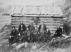 Seven light-skinned men and one darker-skinned man, alternatively sitting and standing, wear coats and bow ties in front of the side wall of a wooden structure