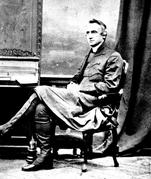 Light-skinned man wearing an open coat over a knee-length tunic and a tall white collar and knee-high boots, sits with his hands clasped in his lap, left leg crossed over, whole body shifted slightly and face looking to the left