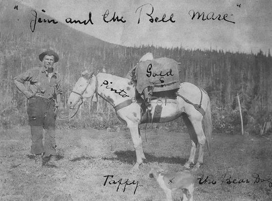 Young man wearing a brimmed hat, a button-up shirt and pants held up by suspenders, smiles with a pipe in his mouth a hand on his hip and holding a rope connected to a pale horse carrying a large bundle of gold wrapped in cloth.