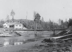 A light-coloured house by a shallow body of water. There is a bridge made of wooden poles over the body of water. 