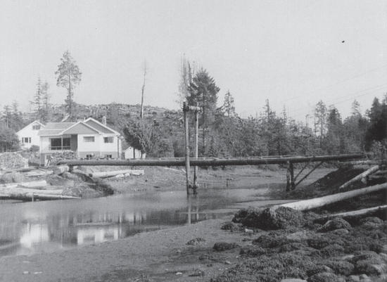A light-coloured house by a shallow body of water. There is a bridge made of wooden poles over the body of water. 