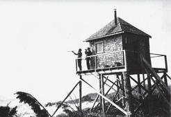 Two people standing outside a wooden lookout, looking into the distance, as seen from below. 