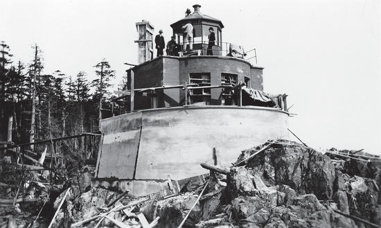 About four people stand on a short lighthouse built on rocks, as seen from below. 