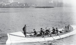 About seven people sit and paddle within a boat on a body of water. One person stands at the front of the boat watching them.