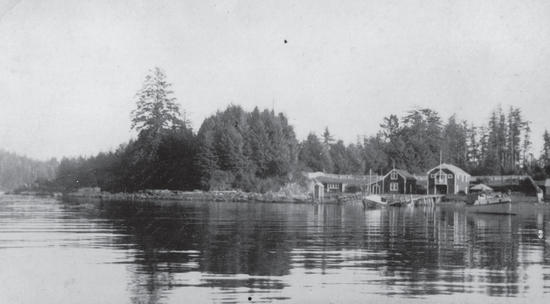A boat at a dock as seen from a body of water. Behind the dock are buildings followed by forest. 
