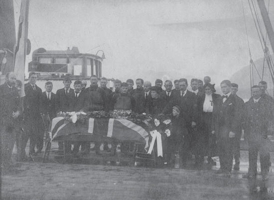 Over 20 people stand posed in formal attire around a casket covered in a flag and flowers. Behind them is a boat. 