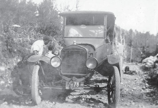 Two people outside an old car on a dirt road. One person sits within the car.