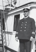 A man with a moustache and a captain's uniform standing on a boat while smiling at the camera. 