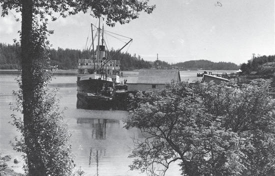A large sailboat docked by a building. Lining the body of water it is on are pieces on land with forests.  