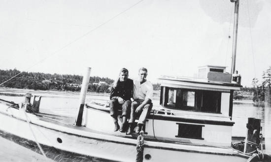 Two people sit on a small, light-coloured boat. The person on the right has an arm around the shoulders of the person on the left. 