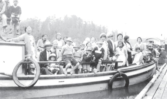 More than 20 people, mostly children, stand and sit on a small, light-coloured boat by a dock while posed for the camera.