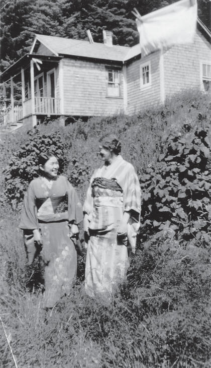 Two women stand in tall grass wearing yukatas while smiling at each other. They stand in front of a house. 