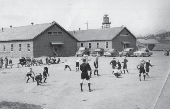 A group of children playing on dirt as another group of children watch while sitting on benches. Behind them are parked cars and buildings. 