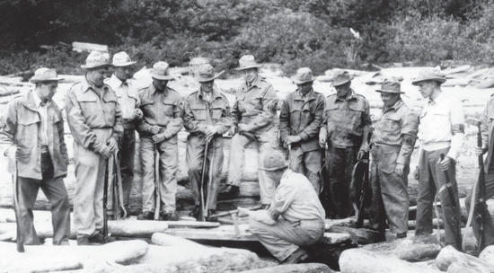 About 10 people in hats and uniforms stand around a person sitting on the ground. They all hold guns. 