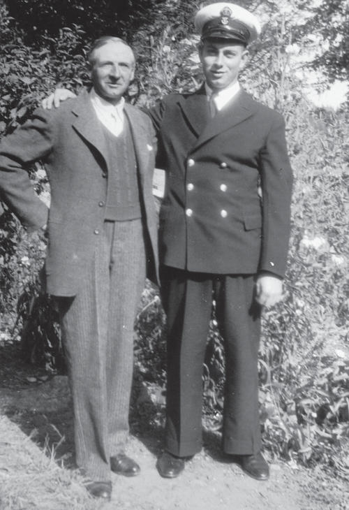Two men stand in front of foliage. The man on the right wears a uniform and his arm is on the shoulder of the other man who wears a sweater vest and a coat. They both look into the camera. 