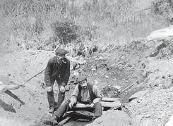A man sits above a small, shallow body of water with a pan. Above him, a man crouches by his side with a pipe in his mouth. They both look at the camera. 