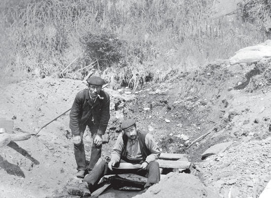 A man sits above a small, shallow body of water with a pan. Above him, a man crouches by his side with a pipe in his mouth. They both look at the camera. 