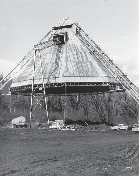 A large, cone-shaped structure suspended above the ground. Vehicles and a building sit under the structure.
