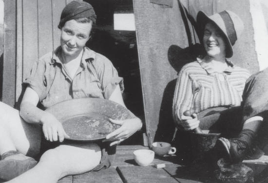 A young woman sits on a wooden floor while holding a pan. She smiles at the camera. Another young woman sits beside her with her legs crossed while grinning at the first woman. 