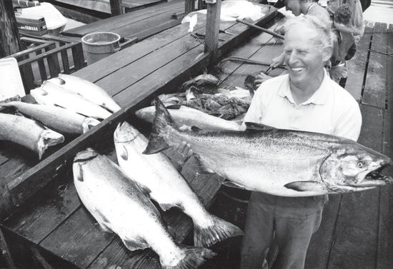 An older man smiling while holding a large fish. Beside him, more large fish lie on a table. 