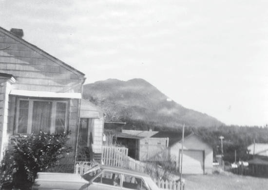 A collection of buildings, a car is parked in front of one of them. Behind them, in the distance, is a large hill. 