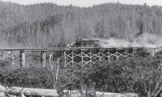 A truck carrying a pile of logs travels across a wooden bridge. Behind the truck is a forest. 
