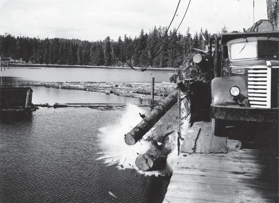 A truck parked on a dock. Logs fall from the truck into the water below. Further back, more logs are floating on the water. 
