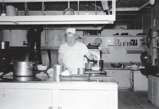 A person looks down, focusing on their work within a kitchen filled with pots, pans, and other cooking tools. 