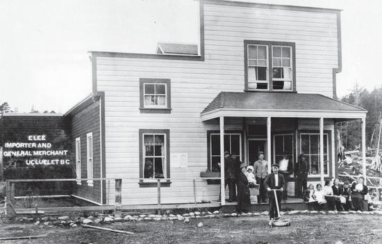 A group of more than 10 people sit and stand in front of a light-coloured building. 
