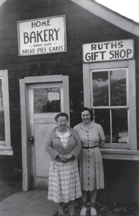 Two women in dresses smile for the camera in front of a storefront. Text: Home Bakery, Home made Bread, Pies, Cakes, Ruths Gift Shop