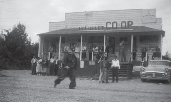 A group of people stand and sit in front of a store. A car with an opened door is in front of the store as well. The front of the store says the following: Ucluelet Co-op