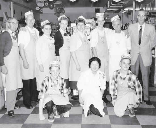Seven people in hats and aprons stand posed for the camera. A man in a suit and glasses stands beside them. In front of them are three more people in aprons, kneeling on the ground. They are within a store. 