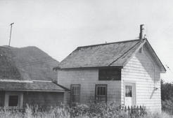 Two buildings beside each other, one lighter and one darker. Behind them are tree-covered hills. 