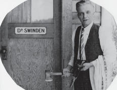 A man in a waistcoat and tie poses with an axe and scissors in front of a door. The text on the door says the following: Dr Swinden
