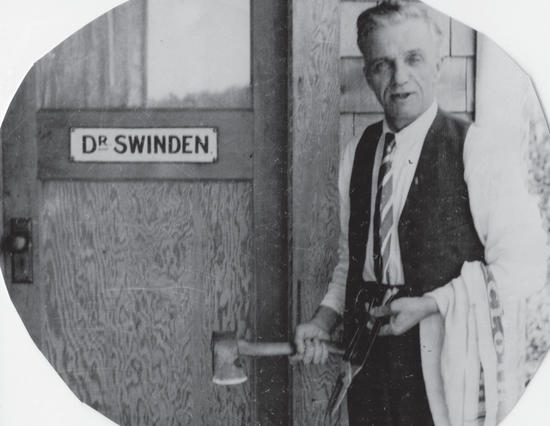 A man in a waistcoat and tie poses with an axe and scissors in front of a door. The text on the door says the following: Dr Swinden
